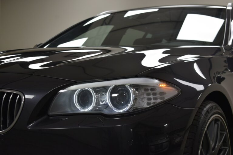 Close-up of the front left side of a dark-colored car, showing the headlights and part of the grille. The car's surface reflects the overhead lights.