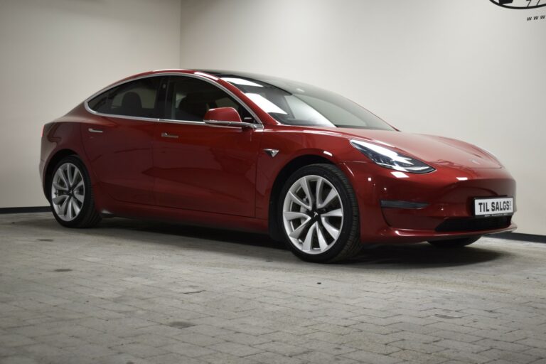 A red Tesla Model 3 is parked indoors on a tiled floor. A sign on the front of the car reads "Til Salgs," indicating it is for sale.