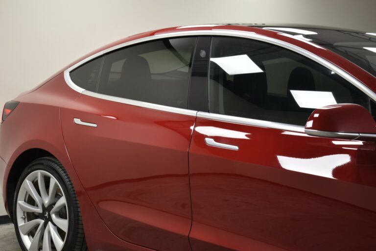 Side view of a red car with tinted windows and shiny silver trim. The car is parked indoors, reflecting ceiling lights on its surface.