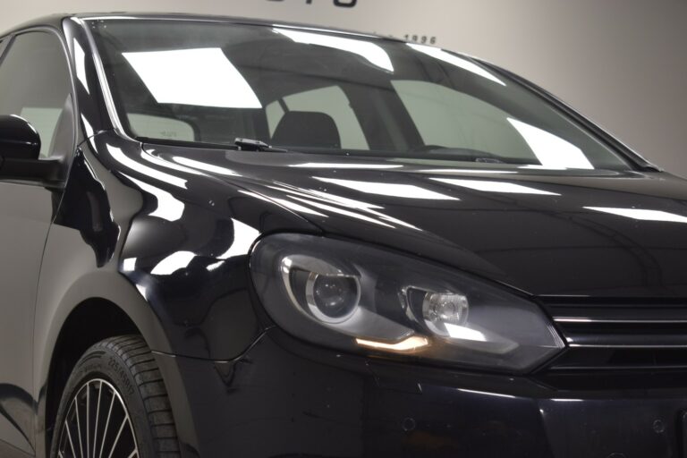 Close-up of a black car's front left side, highlighting the headlight and part of the grille. The setting is indoors with reflective lighting on the car's surface.