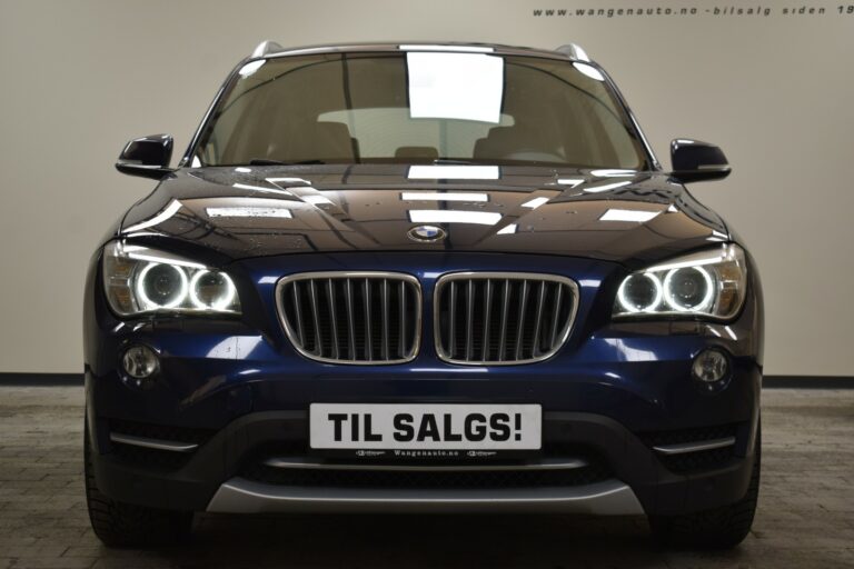 Front view of a dark blue BMW with headlights on. A sign on the bumper reads "Til salgs!" indicating it is for sale.