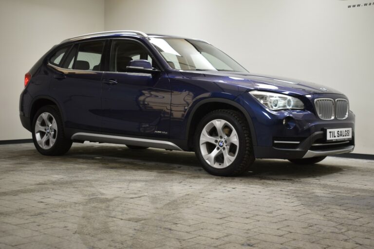 A dark blue BMW X1 parked indoors on a tiled floor, viewed from the side. A "Til Salgs" sign is visible.