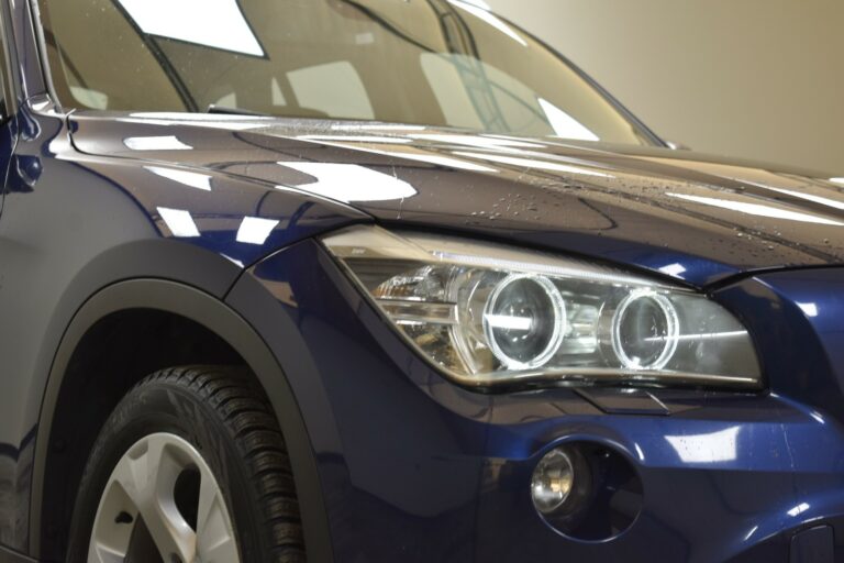 Close-up view of a blue car's front left side, featuring a headlight, part of the wheel, and reflections on the glossy surface.