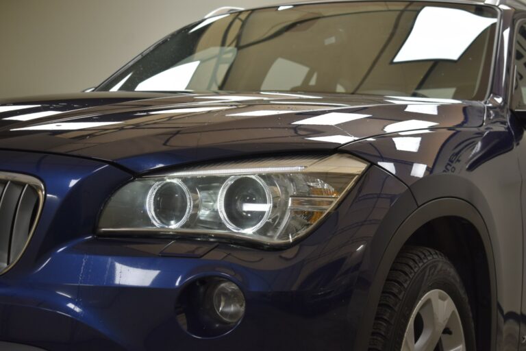 Close-up view of the front left side of a blue car, focusing on the headlight and part of the hood. The background shows reflections of lights on the car's surface.