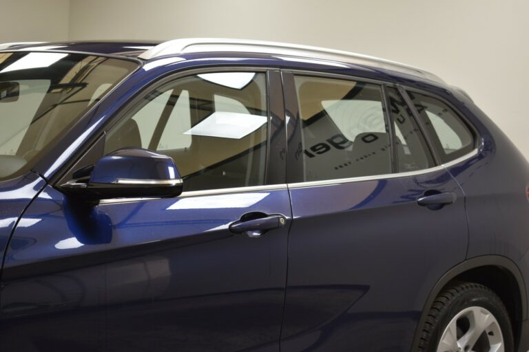 A close-up of a blue SUV from a side angle, showing the side windows, door, and mirror, with part of the interior visible through the glass.