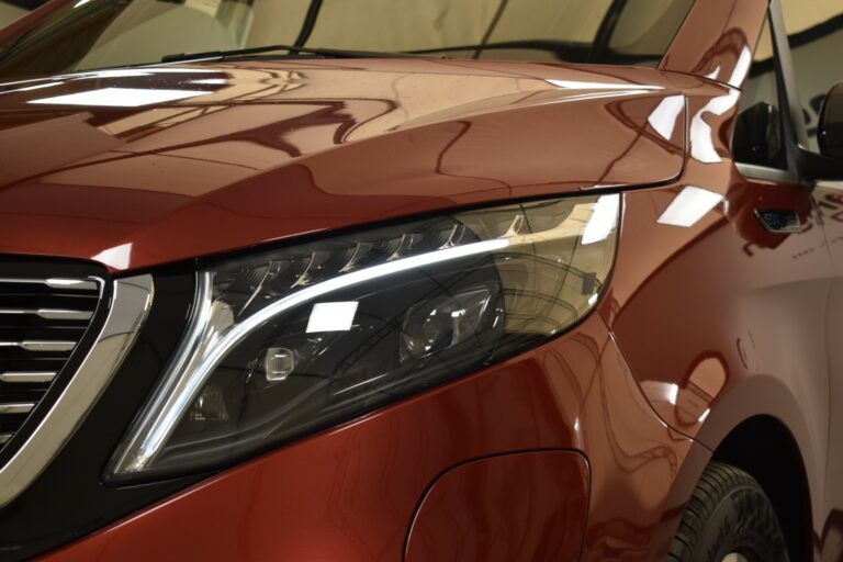 Close-up of a red vehicle's front headlight and grill, showing sleek lines and reflective surfaces.