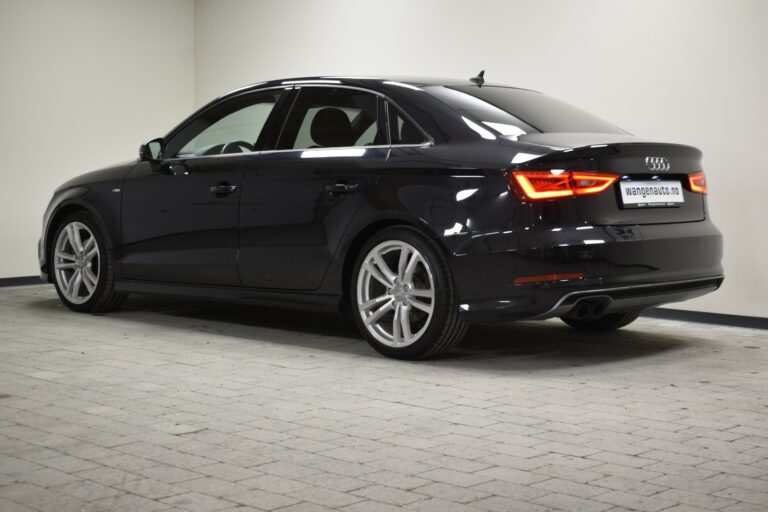 A black Audi sedan with illuminated rear lights, parked in a well-lit indoor space, viewed from the rear right side.