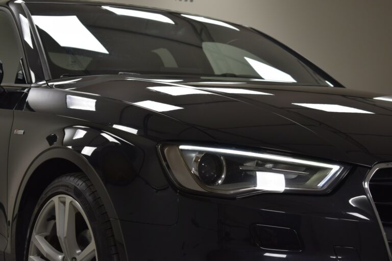 Close-up of the front end of a black sedan with illuminated headlights and visible reflections on the hood and windshield.