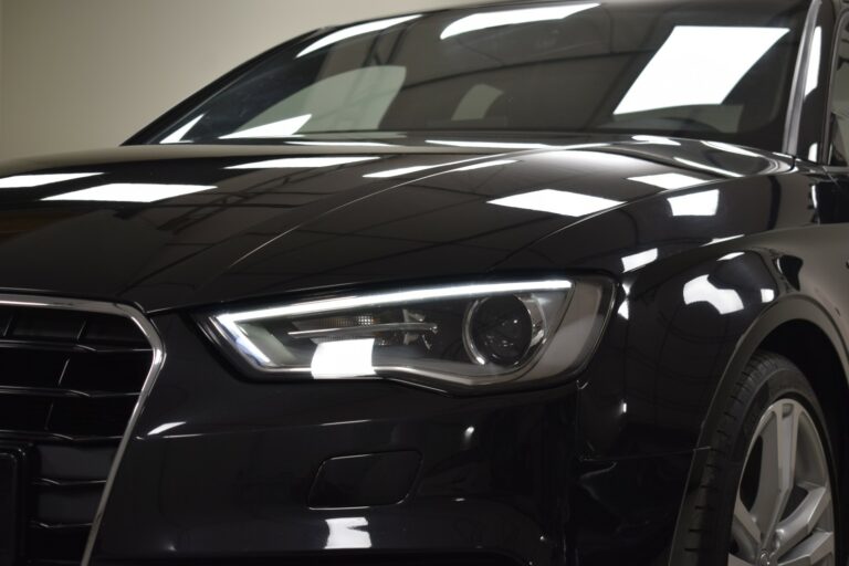 Close-up of the front left side of a black car, showing a sleek headlight and part of the hood and grille against a dimly lit background.