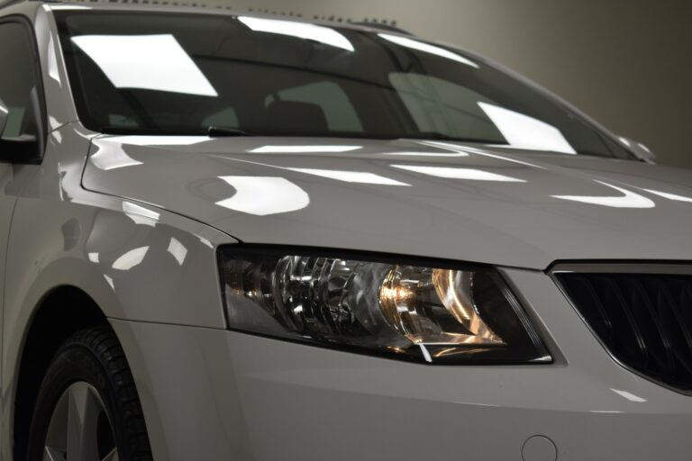 Close-up of the front left side of a white car, highlighting the headlight and part of the hood in a well-lit indoor setting.