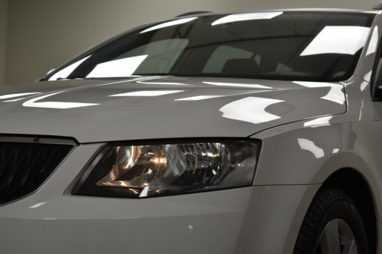 Close-up of a white car's front section showing the headlight and part of the hood. The headlight is illuminated, and reflections are visible on the car's surface.