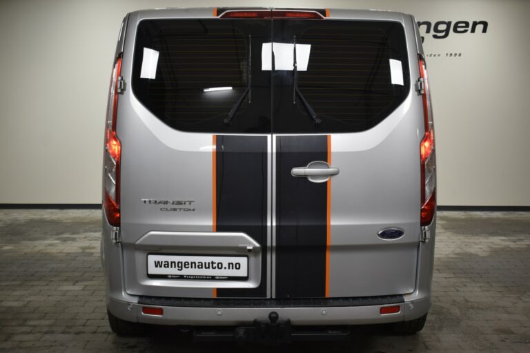 Rear view of a silver Ford Transit Custom van with black and orange stripes, parked indoors in front of a sign that reads "wangenauto.no".