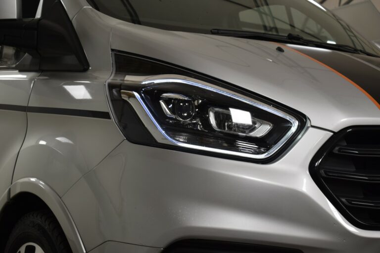 Close-up of a silver van's front headlight and part of the grille, featuring modern design elements and reflective lighting.