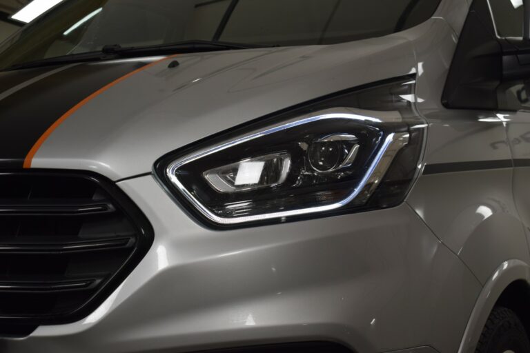 Close-up of a silver van's front end, highlighting the headlight with a distinctive LED strip and orange racing stripe on the hood.