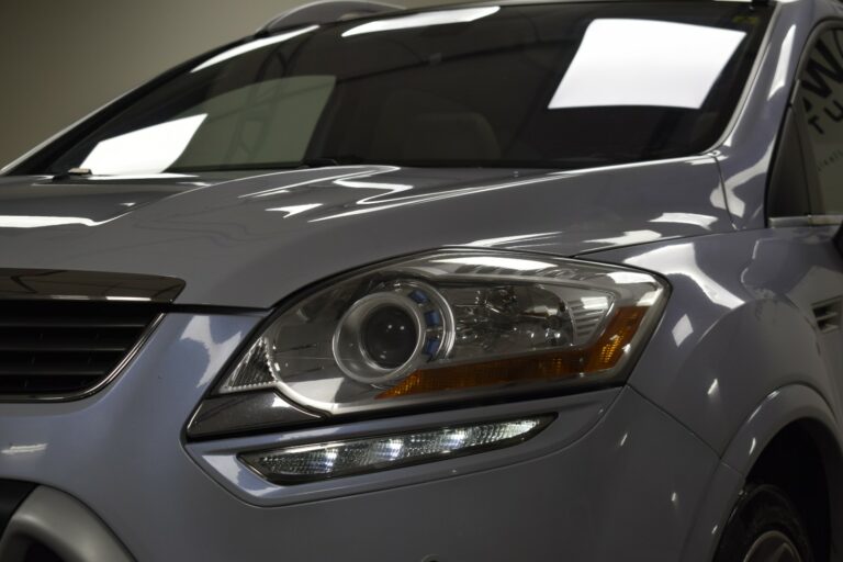 Close-up of a car's front headlight and part of the hood, showing reflective details and a sleek design.