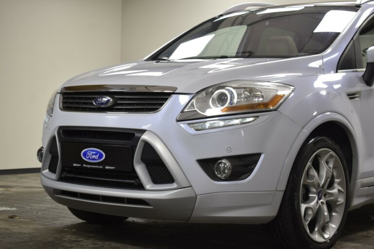 A silver Ford SUV is parked indoors, showing a close-up of the front end, grille, and headlights. The Ford logo is visible on the grille and front license plate.